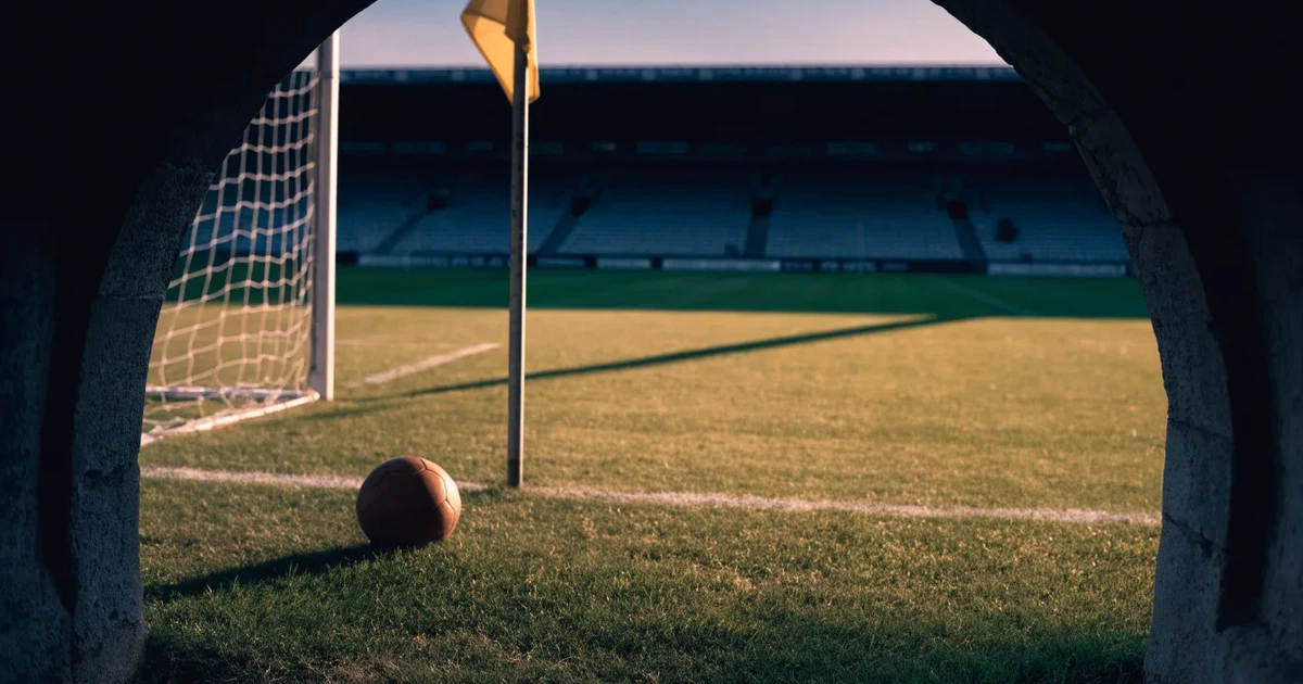 Blick durch ein Stadiontor auf einen grünen Rasen im Abendlicht, ein Ball ruht an der Eckfahne, die Tribünen sind leer.