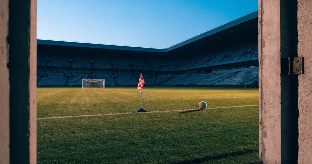 Blick durch ein Stadiontor auf einen grünen Rasen im Abendlicht, ein Ball liegt an der Eckfahne, die Ränge sind leer.