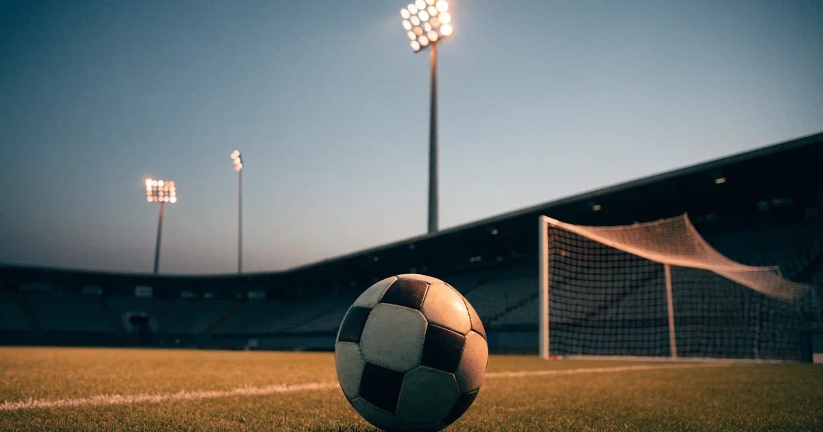 Ein Fußball liegt auf dem Rasen eines leeren Stadions bei Flutlicht, im Hintergrund unscharfe Tribünen ohne erkennbare Beschriftung.