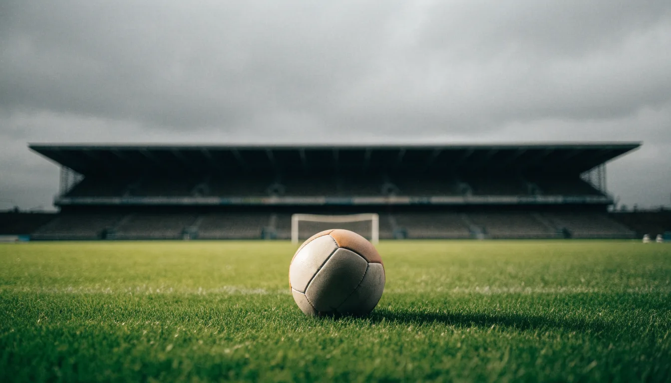 Ein Fußball auf einem Rasen, im Hintergrund ein leeres Stadion bei bewölktem Himmel.