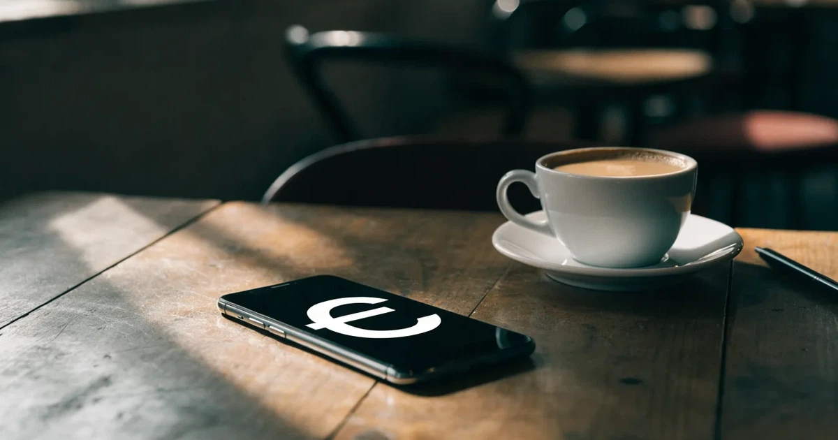 Ein Smartphone liegt mit dunklem Display auf einem Café-Tisch neben einer Tasse Kaffee und einem Kugelschreiber.