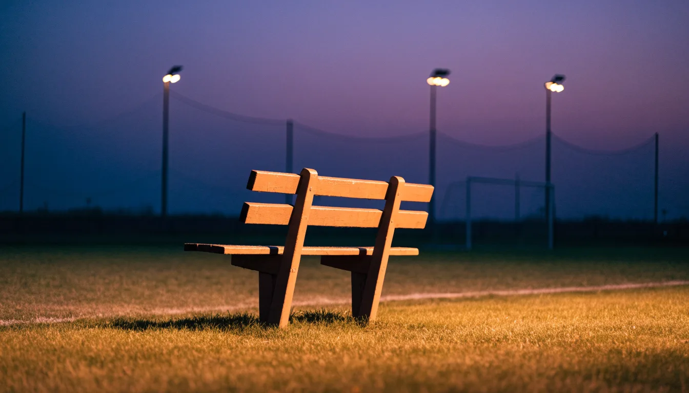 Eine leere Parkbank am Rand eines Fußballfelds in der Abenddämmerung, im Hintergrund ragen dunkle Flutlichtmasten in den Himmel.