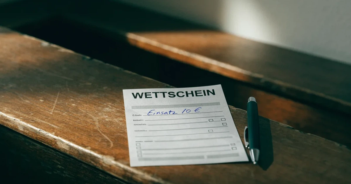 Ein leerer Wettschein aus Papier liegt neben einem Stift auf einer hölzernen Theke, weiches Licht fällt von der Seite ein.