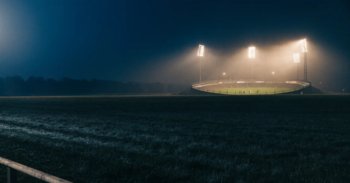 Ein Stadion unter Flutlicht, aus großer Entfernung über eine dunkle Wiese fotografiert, am Himmel zieht Nebel auf.