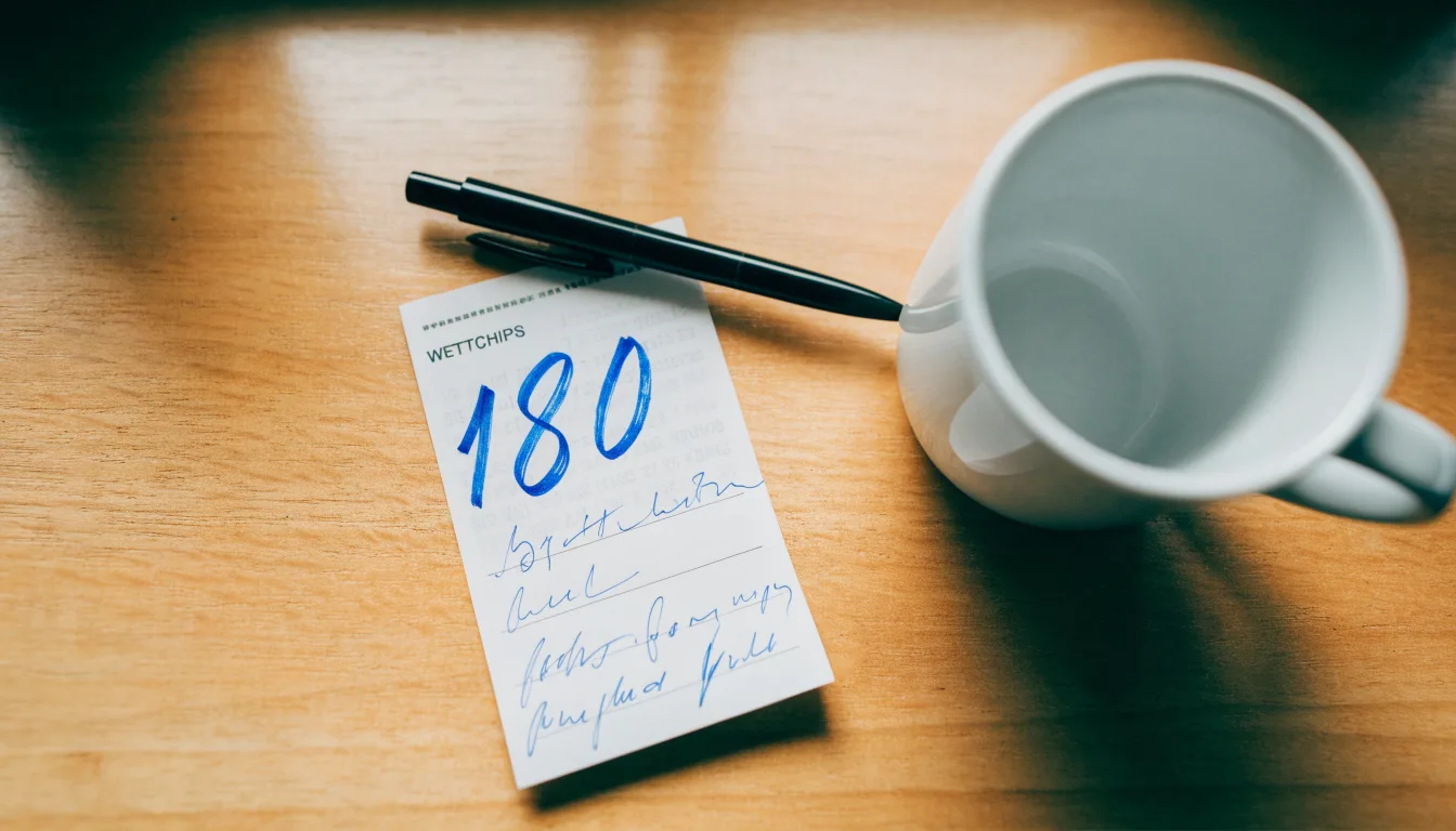 Ein leerer Wettschein aus Papier liegt neben einem Kugelschreiber und einer Tasse auf einem hellen Holztisch.