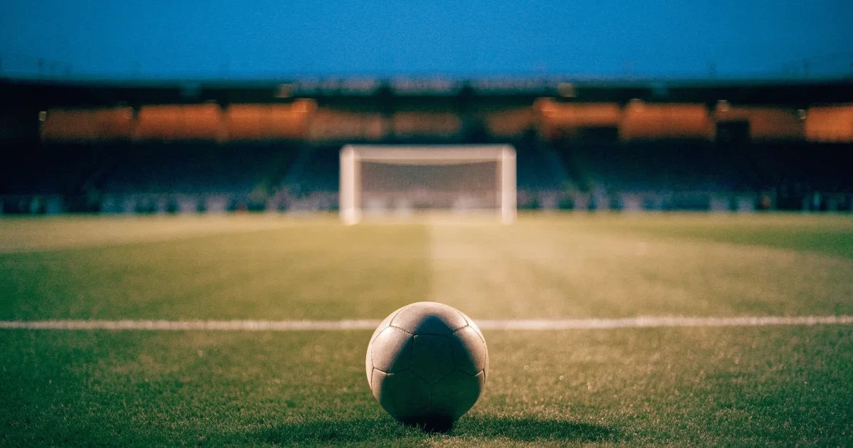 Ein Fußballplatz mit frisch gemähtem Rasen im Abendlicht, ein Ball liegt im Strafraum, im Hintergrund unscharf eine leere Tribüne.
