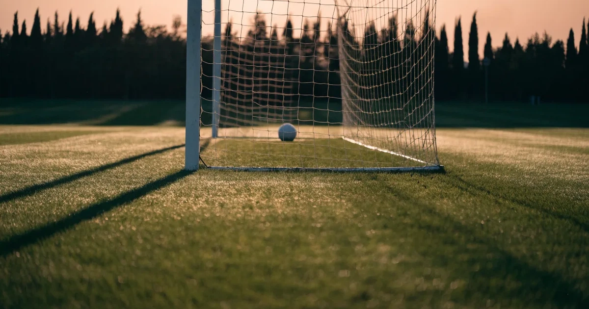 Ein Fußballtor mit Netz auf einem leeren Platz im Abendlicht, der Rasen frisch gemäht, im Hintergrund Zypressen.