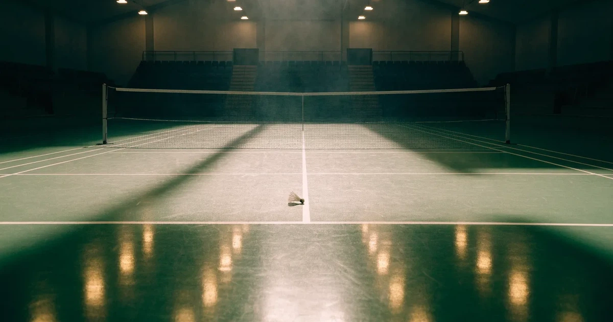 Eine leere Badmintonhalle mit gespanntem Netz und hellen Hallenlichtern, ein Federball liegt auf dem Hallenboden.