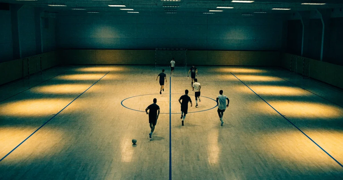 Eine leere Sporthalle mit hellem Hallenboden, kleinen Toren an beiden Enden und blauen Linien, Licht fällt von oben.