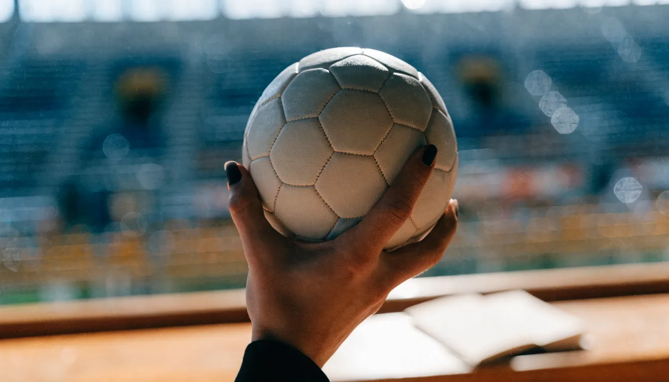 Eine Hand hält einen Handball, im Hintergrund eine unscharfe Tribüne, daneben liegt ein Notizblock auf einer Bank.