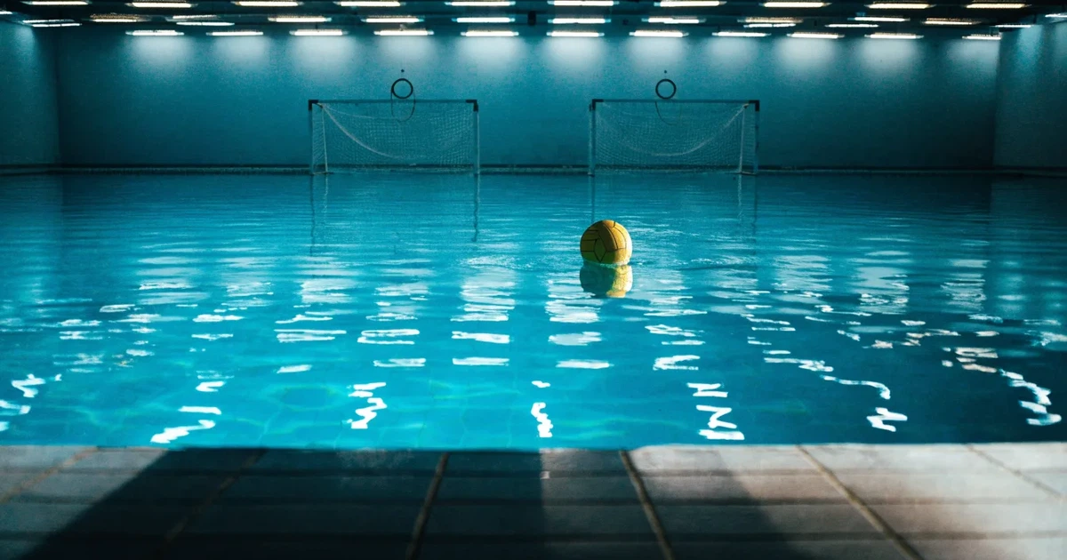 Ein Schwimmbecken mit türkisfarbenem Wasser und zwei schwimmenden Torrahmen am gegenüberliegenden Beckenrand, menschenleer unter Hallenlicht.
