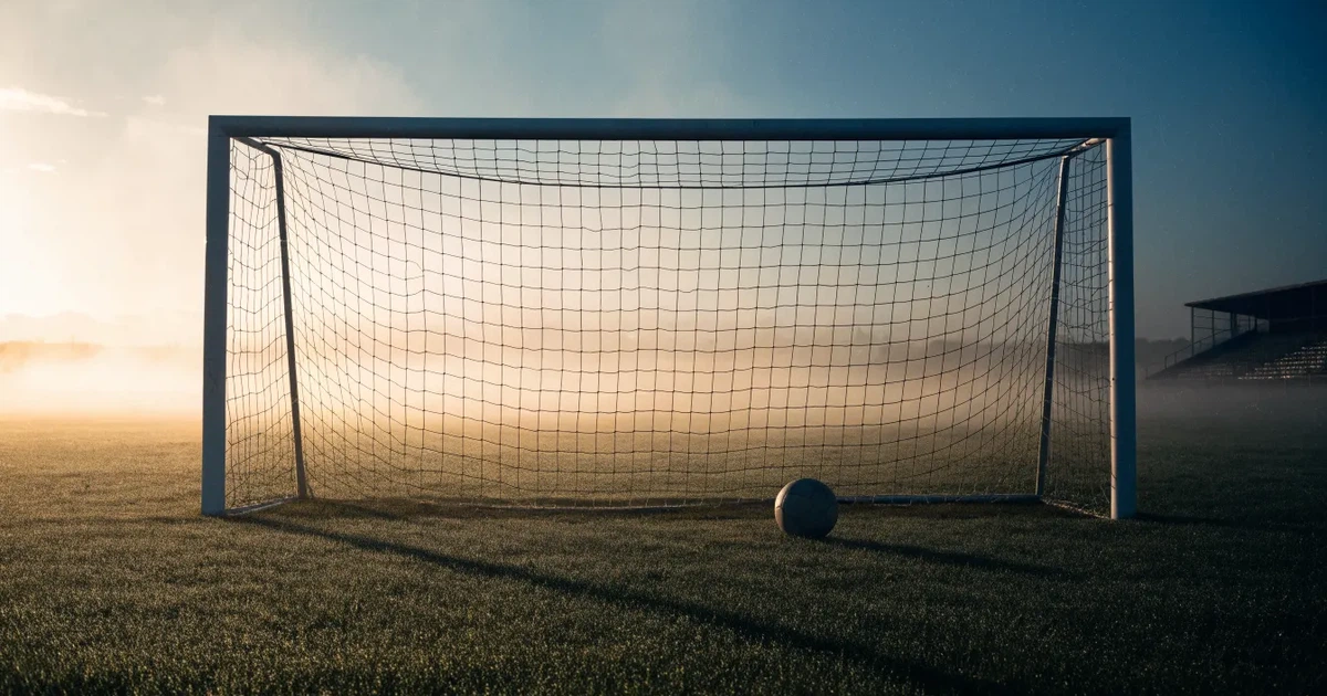 Ein Fußballtor mit Netz steht im Morgennebel auf einem leeren Rasenplatz, im Hintergrund die Silhouette einer kleinen Tribüne.