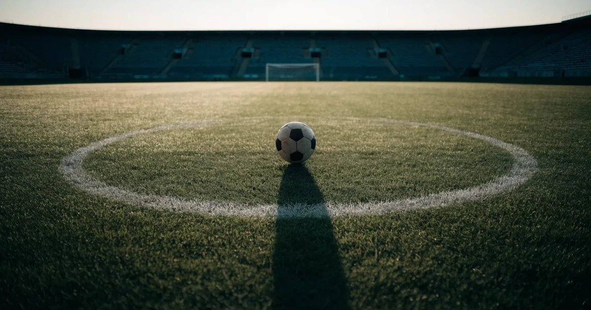 Ein Fußball liegt auf dem Anstoßpunkt eines Rasenplatzes, dahinter leere Tribünen im Licht des frühen Morgens.
