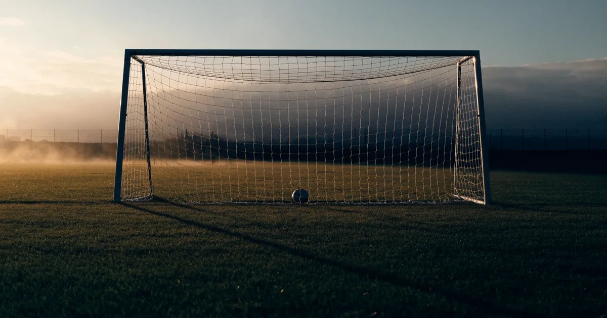 Ein Fußballtor mit Netz im Gegenlicht auf einem leeren Platz am Abend, davor ein Ball auf dem Rasen.